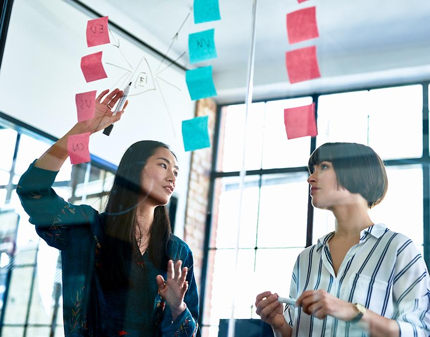 Two women looking at post it notes on glass board